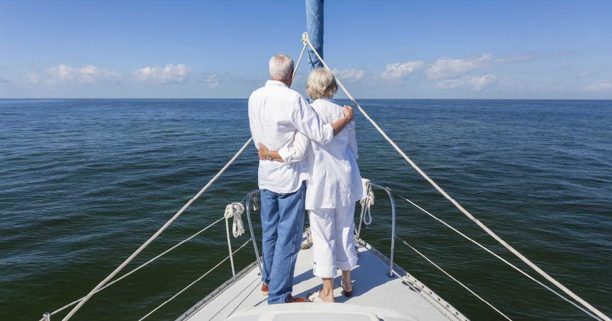 A couple that has achieved their retirement dream is standing on a sailboat looking into the distance.